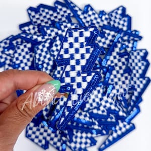 May include: Blue and white checkered lightning bolt patch with blue sequin trim. The patch has a detailed checkered pattern and is held in a hand. Many similar patches are in the background.