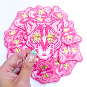 May include: Pink embroidered tiger face patch with gold star eyes. The patches are arranged in a circular pattern. The patch is a vibrant pink with gold glitter accents. The patches are suitable for clothing and accessories.