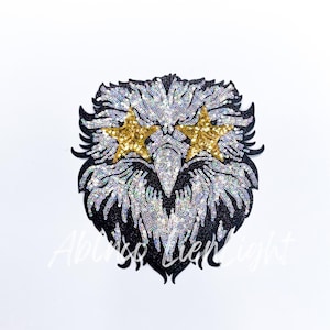 May include: A black and silver sequined eagle head with gold star-shaped eyes. The eagle's beak is black and the feathers are silver with a black outline.