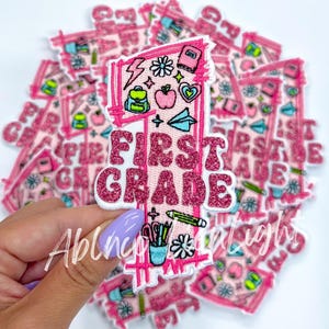 May include: Pink and white iron-on patch with the text "First Grade" in pink glitter letters. The patch features a number one with school supplies and other designs.