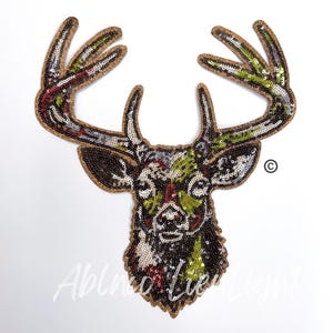 May include: A colorful sequined deer head applique with brown, green, white, and red sequins. The deer has large antlers and a detailed face.