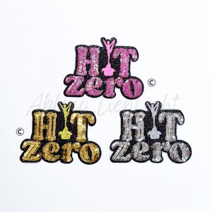 May include: Three sequined patches with the words "HIT zero" in pink, gold, and silver. Each patch has a black border and a small trophy graphic. The patches are on a white background.