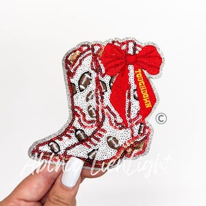 May include: A sequined patch in the shape of a cowboy boot, featuring a white base with brown footballs. A red bow with the word "TOUCHDOWN" in gold lettering is attached. The boot has a silver glitter border.