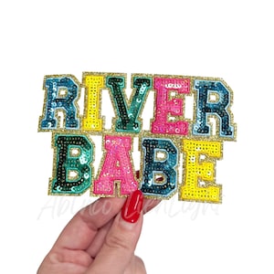 May include: A colorful sequined patch with the words "RIVER BABE" in a collegiate font. The letters are in shades of blue, yellow, pink, and green, outlined with gold glitter. The patch is held up by a hand with red nail polish.