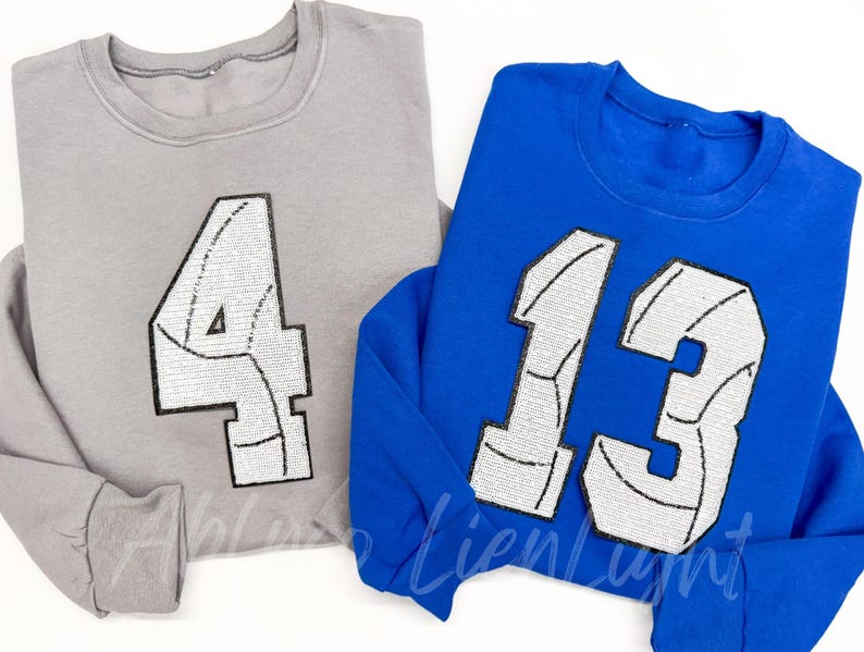 K&ouml;nnte beinhalten: Zwei graue und blaue Sweatshirts mit wei&szlig;en Volleyball-Nummern. Das graue Sweatshirt hat die Nummer "4" und das blaue Sweatshirt die Nummer "13".