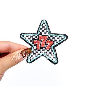 May include: A star-shaped patch with a black and white checkered pattern. The patch features the number "777" in red glitter. The star is outlined in black and has a turquoise border.