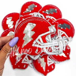 May include: A stack of red felt patches with a silver sequin lightning bolt and white embroidered football helmet design.