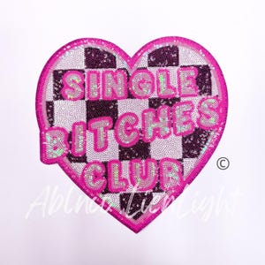 May include: A heart-shaped patch with a checkerboard pattern and the words "SINGLE BITCHES CLUB" in pink, sequined letters. The patch has a pink border and is set against a white background.