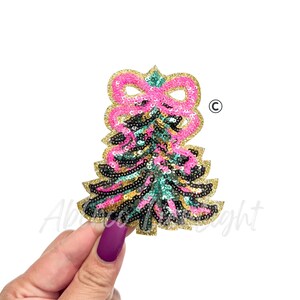 May include: A sequined Christmas tree patch with a pink bow at the top. The tree is decorated with pink, black, and teal sequins, outlined in gold glitter. The patch is held by a hand with a purple painted nail.