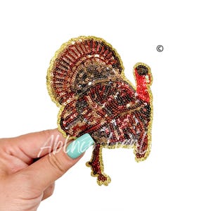 May include: A sequined turkey applique with a red, brown, and gold color scheme. The turkey has a red head and a fanned tail. The applique is held by a hand with a turquoise nail polish.