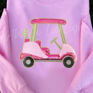 May include: A light pink sweatshirt featuring a golf cart design. The golf cart is embellished with pink sequins and gold accents. The text "Able to Lie Light" is faintly visible on the front.
