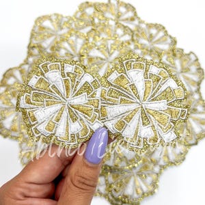 May include: A pile of gold and white glitter embroidered fabric flower appliques. The appliques are about 2 inches in diameter and have a geometric design.