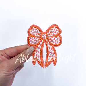 May include: Embroidered bow applique in coral and white gingham pattern. The bow has a coral outline and is held by a hand. The text "Ablinca Ley Light" is visible on the image.