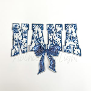 May include: A white and blue floral patterned iron-on patch with the word "NANA" in large letters and a blue sequined bow.