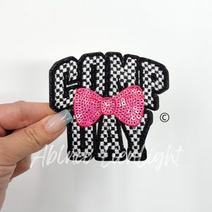May include: Black and white checkered fabric patch with the word "Camp" and a pink sequined bow. The word "Day" is below the bow.