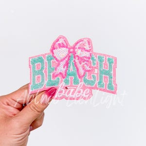 May include: A pink and green iron-on patch with the word "BEACH" in large, glittery letters. A pink bow with glitter is centered above the word.