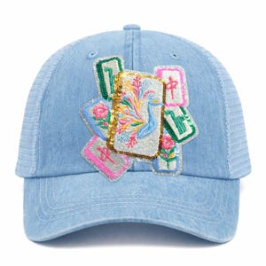 May include: Light blue denim baseball cap with a white mesh back. The cap features embroidered patches with floral and peacock designs, accented with sequins. The patches are in shades of pink, green, and gold.