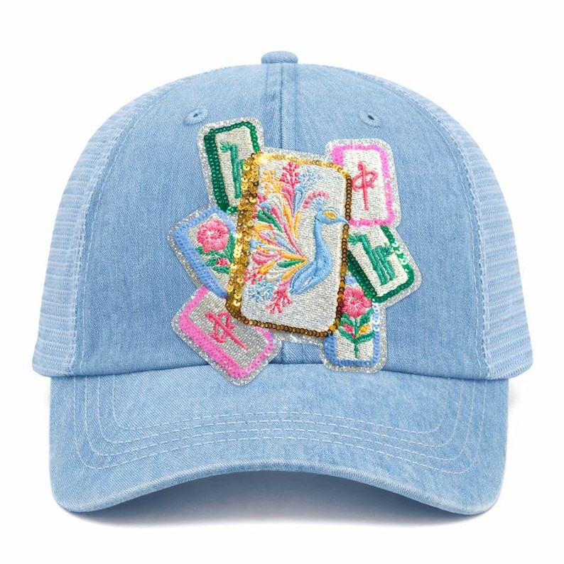 May include: Light blue denim baseball cap with a white mesh back. The cap features embroidered patches with floral and peacock designs, accented with sequins. The patches are in shades of pink, green, and gold.