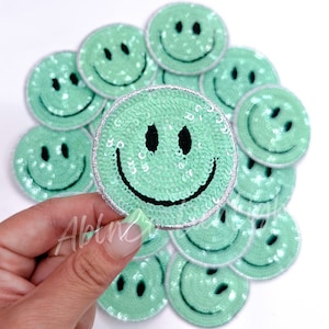 May include: A set of 10 green sequin smiley face iron-on patches. The patches are approximately 2 inches in diameter and have a silver trim.