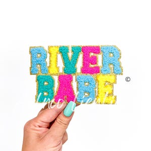 May include: A colorful felt letter banner that reads "River Babe" with gold glitter outlining each letter. The letters are in shades of blue, yellow, pink, and turquoise.