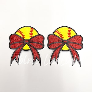 May include: Two softball-themed embellishments. Each features a yellow softball with red stitching, topped with a red glitter bow with black trim. The background is white.