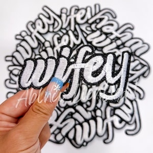 May include: A pile of black and white iron-on patches with the word "wifey" in white embroidery. The patches are arranged in a circle with the word "wifey" in the center.