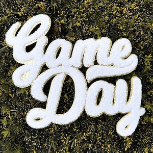 May include: White fluffy letters spelling "Game Day" with gold glitter outlining the letters. The letters are on a green background.