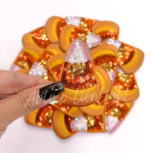 May include: A pile of orange, yellow, and white candy corn shaped iron-on patches with sequins. The patches are approximately 2 inches in size.