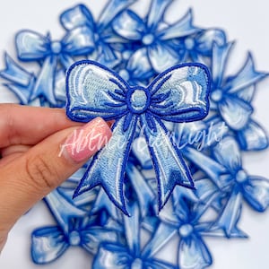 May include: A close-up of a blue satin bow with a white center. The bow is embroidered and has a slightly iridescent sheen. The bow is part of a set of multiple bows.