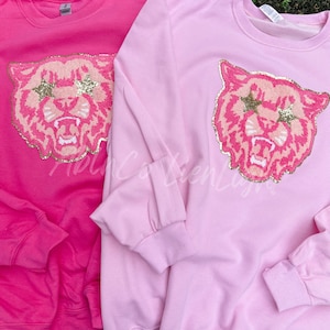 May include: Two sweatshirts with a pink tiger face with gold glitter stars on the front. One sweatshirt is pink and the other is hot pink.
