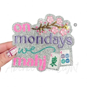 May include: Glittery patch with the text "on mondays we mahj". The patch features pink cherry blossoms, a branch with gold sequins, and embroidered mahjong tiles. The patch is held by a hand.