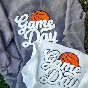 May include: Two sweatshirts with the text "Game Day" in white with a basketball graphic. The sweatshirt on the left is gray and the sweatshirt on the right is white.