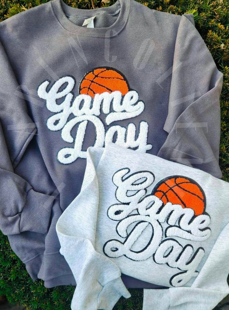 May include: Two sweatshirts with the text "Game Day" in white with a basketball graphic. The sweatshirt on the left is gray and the sweatshirt on the right is white.