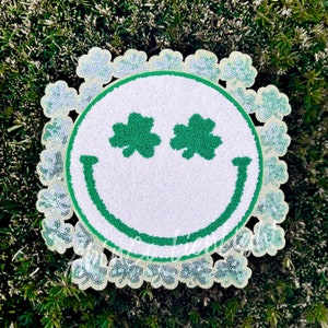 May include: A white smiley face patch with green shamrock eyes surrounded by a border of silver glitter shamrocks.