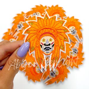 May include: A close-up of a pile of orange and white embroidered patches featuring a stylized face with a headdress. The face has a white outline and features a gray nose, mouth, and eyes with gold stars.
