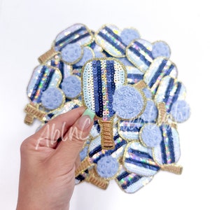 May include: A pile of blue and gold sequin iron-on patches shaped like a paddle and ball. The patches have a fuzzy blue ball and a blue and white striped paddle with gold trim.
