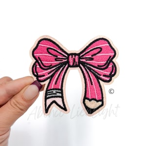 May include: Pink and black embroidered iron-on patch of a bow with a pencil for the bow's tail. The pencil has a pink eraser.