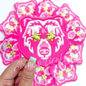 May include: Pink felt iron-on patch with a white bear face design. The bear has large, white, star-shaped eyes with gold glitter accents. The patch is surrounded by other similar patches.