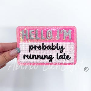 May include: Pink sparkly iron-on patch with the text "HELLO I'M probably running late" in black.