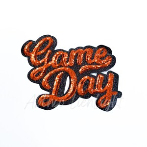 May include: A sequined patch with the words "Game Day" in orange script lettering. The letters are outlined in black glitter and are on a white background. The patch is likely for apparel or accessories.