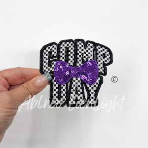 May include: A black and white checkered iron-on patch with the text "Camp Day" and a purple sequin bow.