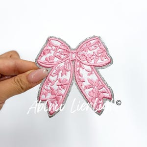 May include: A pink and silver embroidered bow with a floral design. The bow is on a white background.