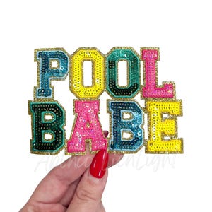 May include: A colorful sequined patch that spells out "POOL BABE" in block letters. The letters are in various colors, including teal, yellow, pink, and green, with gold glitter borders. The patch is held by a hand with red nail polish.