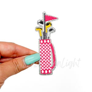 May include: A pink and white checkered golf bag with a red flag and yellow golf clubs. The bag has white polka dots.