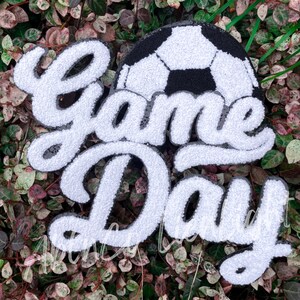 May include: White and black felt applique with the words "Game Day" in a cursive font. A black and white soccer ball is above the words.