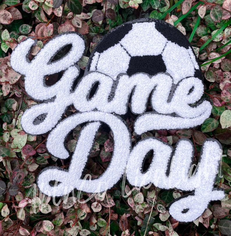 May include: White and black felt applique with the words "Game Day" in a cursive font. A black and white soccer ball is above the words.