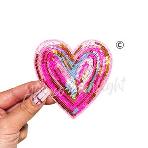 Puede incluir: Un parche de lentejuelas en forma de coraz&oacute;n en tonos rosa, dorado y azul claro. El parche se sostiene sobre un fondo blanco. Las lentejuelas est&aacute;n dispuestas en c&iacute;rculos conc&eacute;ntricos, creando un efecto texturizado. El parche es probablemente para fines decorativos.