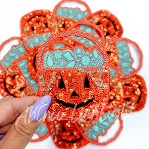 May include: Orange sequined pumpkin-shaped patches with black facial features and a teal top. The patches have a handle and are decorated with sequins. The patches are for Halloween.