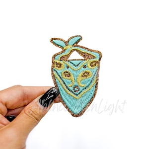 May include: A turquoise blue bandana with gold embroidery, featuring a stylized design of a face with two eyes and a nose. The bandana is tied at the top and has a pointed bottom.