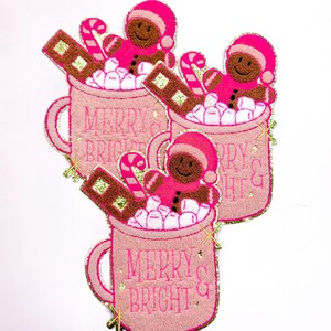 May include: Three pink and gold embroidered patches featuring a mug filled with marshmallows, candy canes, and chocolate. A gingerbread person is in each mug. The text "Merry & Bright" is on each mug.